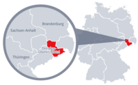 Diese Grafik zeigt eine Karte von Deutschland mit einer detaillierten Ansicht der Region Sachsen-Dresden-Oberes-Elbtal. Sachsen-Dresden-Oberes-Elbtal ist in Rot hervorgehoben und liegt im östlichen Teil Deutschlands. Ein Lupensymbol vergrößert diesen Bereich, um die Lage genauer darzustellen. 